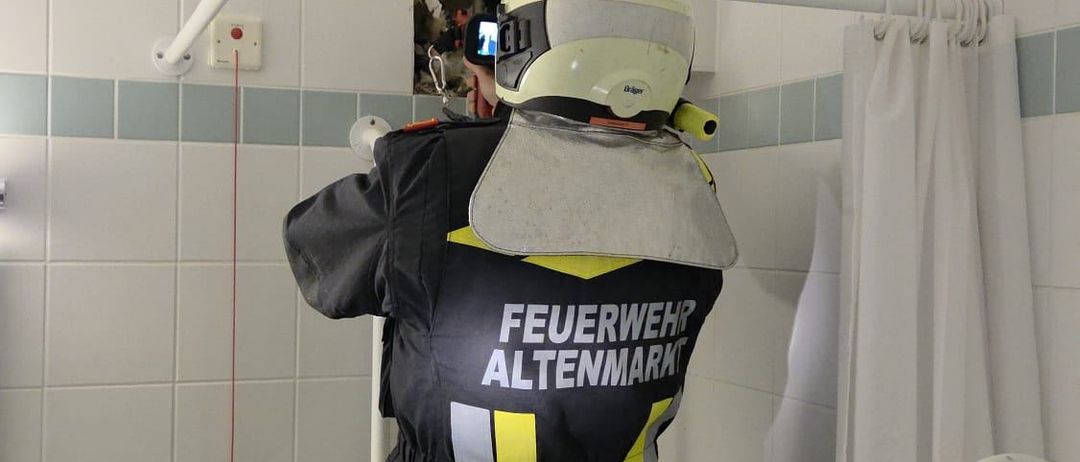 Ein Feuerwehrmann in einem Badezimmer mit einer gebrochenen Wand, der einen Helm und eine reflektierende Weste trägt. Er hält eine Kamera und scheint zu filmen.