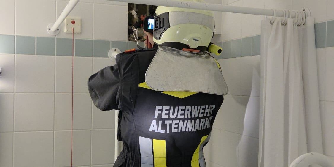 Ein Feuerwehrmann in einem Badezimmer mit einer gebrochenen Wand, der einen Helm und eine reflektierende Weste trägt. Er hält eine Kamera und scheint zu filmen.