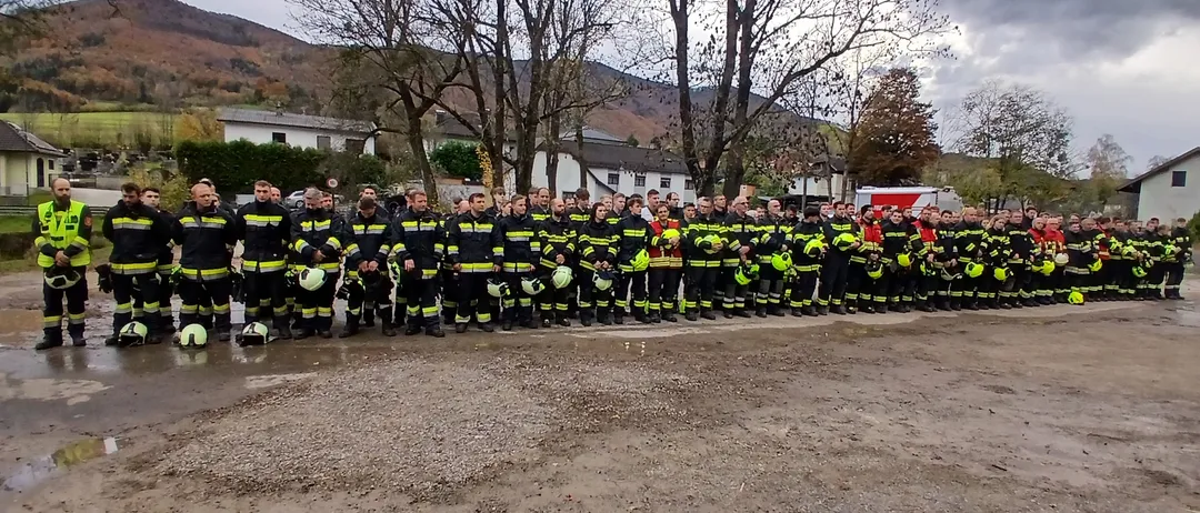 Eine Gruppe von Feuerwehrleuten steht in einer Reihe auf nassem Boden, in voller Ausrüstung. Sie stehen vor Bäumen und Gebäuden, mit Bergen im Hintergrund.