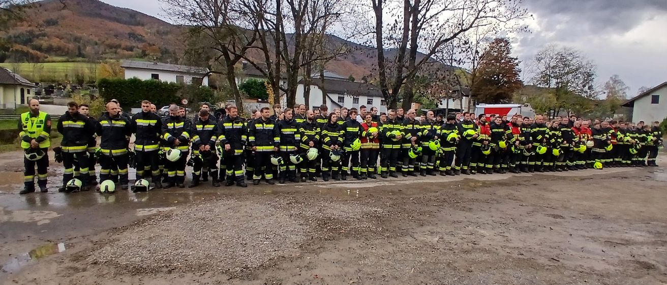 Eine Gruppe von Feuerwehrleuten steht in einer Reihe auf nassem Boden, in voller Ausrüstung. Sie stehen vor Bäumen und Gebäuden, mit Bergen im Hintergrund.