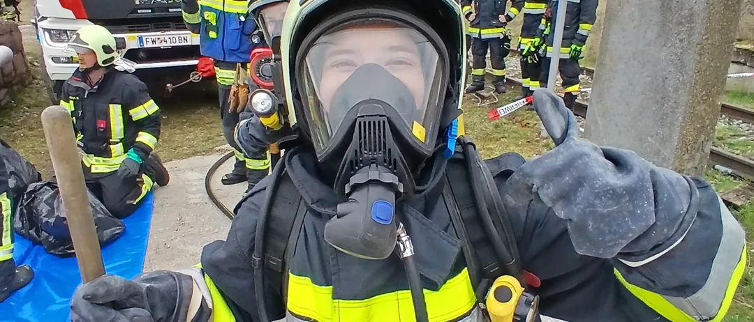 Ein Feuerwehrmann in voller Ausrüstung steht mit einem Stab und gibt ein Daumenhoch, mit Feuerwehrwagen und Kollegen im Hintergrund.
