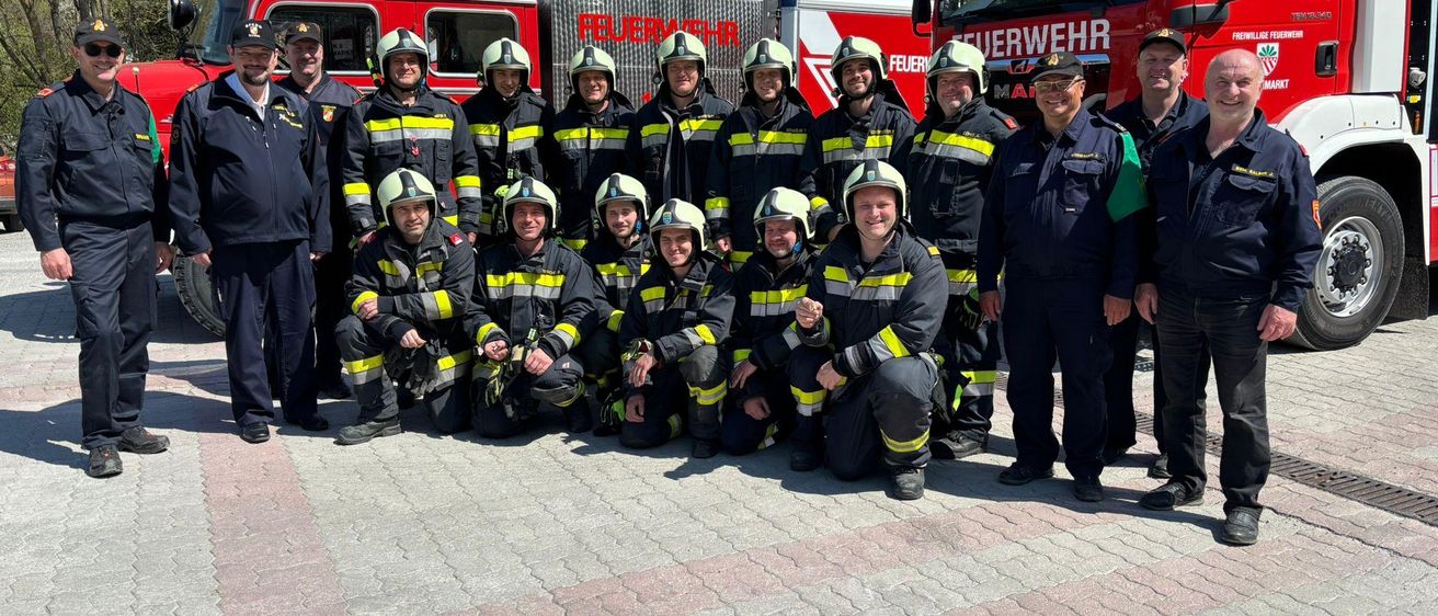 Eine Gruppe von Feuerwehrleuten posiert für ein Foto, trägt Uniformen und Helme. Einige knien, und sie stehen vor einem roten Feuerwehrwagen mit Text.