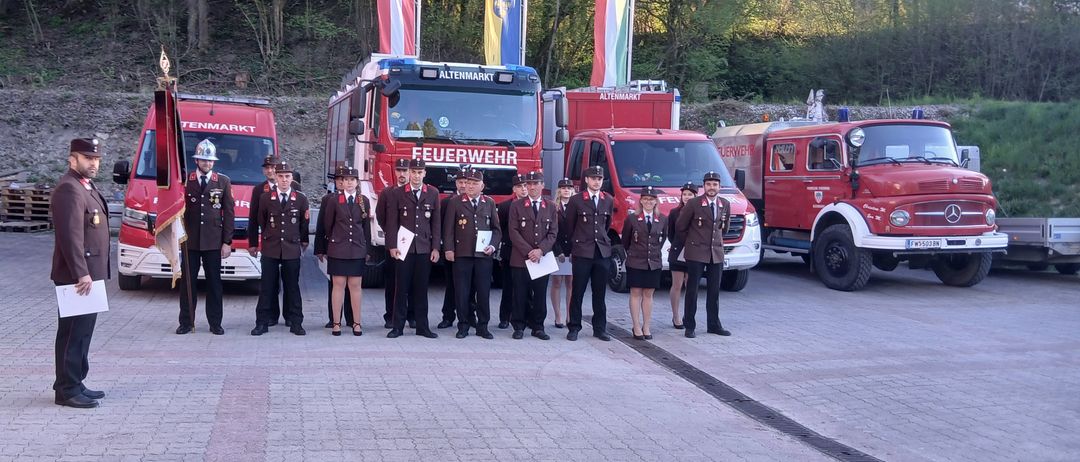 Eine Gruppe Feuerwehrleute in Uniform steht vor roten Feuerwehrfahrzeugen, eines mit dem Wort 'Feuerwehr' darauf. Dahinter befinden sich Fahnen.