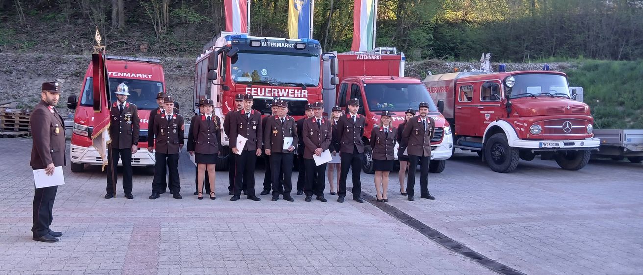 Eine Gruppe Feuerwehrleute in Uniform steht vor roten Feuerwehrfahrzeugen, eines mit dem Wort 'Feuerwehr' darauf. Dahinter befinden sich Fahnen.