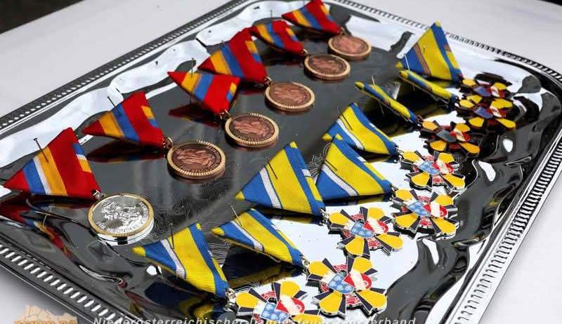 Ein silbernes Tablett zeigt mehrere Medaillen, einschließlich roter, gelber und blauer Bänder mit goldenen Zentren. Die Medaillen sind in einer Reihe angeordnet und reflektieren die Oberfläche des Tabletts.