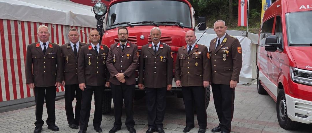 Sieben Männer in Uniform stehen vor einem Feuerwehrauto mit Flaggen und einem nahe gelegenen Van.