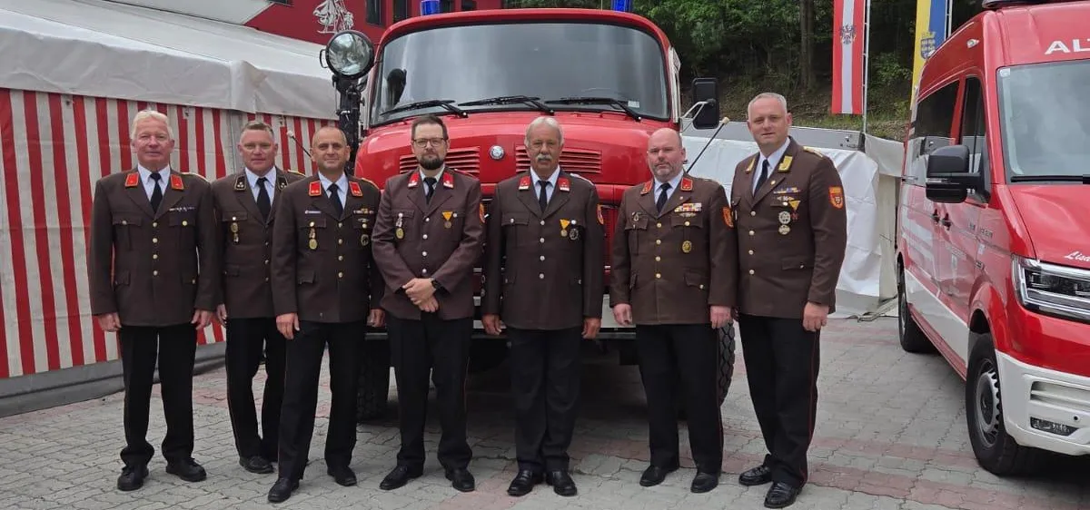Sieben Männer in Uniform stehen vor einem Feuerwehrauto mit Flaggen und einem nahe gelegenen Van.