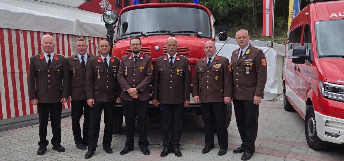 Sieben Männer in Uniform stehen vor einem Feuerwehrauto mit Flaggen und einem nahe gelegenen Van.