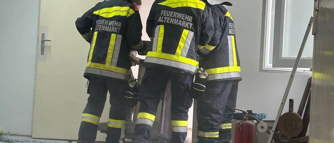 Drei Feuerwehrleute in Schutzkleidung stehen in einem Raum, möglicherweise zur Untersuchung eines Vorfalls. Der Raum hat weiße Wände und eine offene Tür. Trümmer und ein Feuerlöscher liegen auf dem Boden.