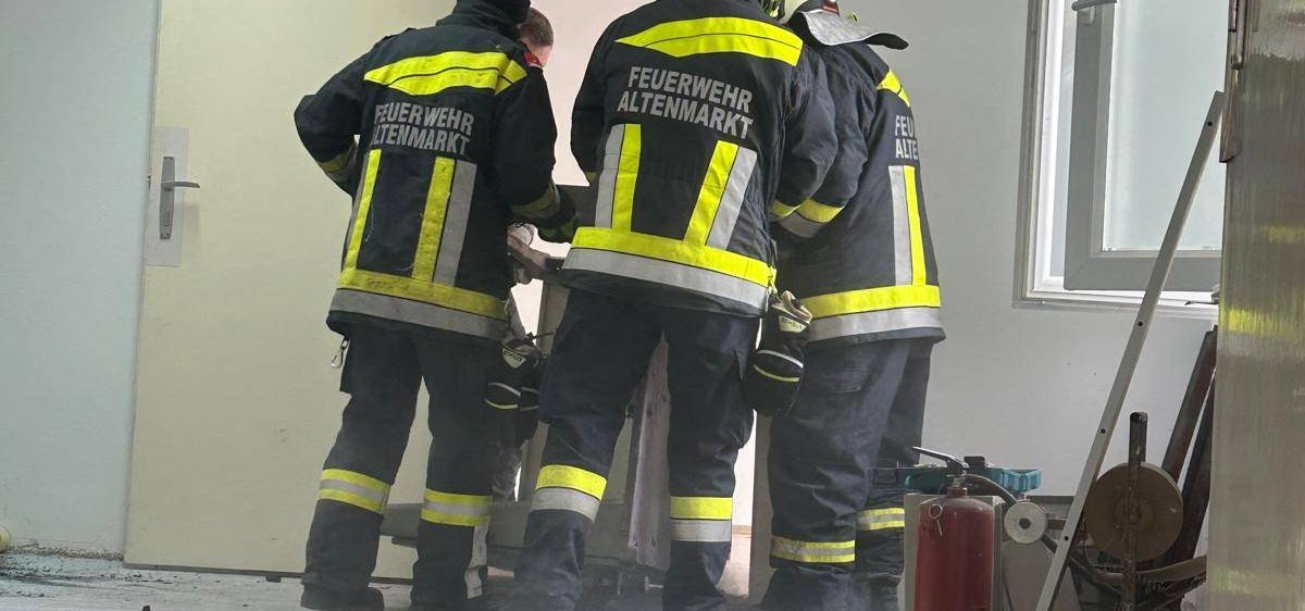 Drei Feuerwehrleute in Schutzkleidung stehen in einem Raum, möglicherweise zur Untersuchung eines Vorfalls. Der Raum hat weiße Wände und eine offene Tür. Trümmer und ein Feuerlöscher liegen auf dem Boden.