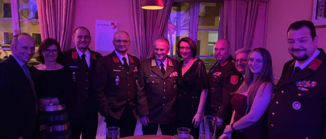 Eine Gruppe von Menschen, einige in Militäruniformen, posiert für ein Foto in einem Raum mit lila Beleuchtung. Es gibt Gläser und ein Tisch mit Getränken und Snacks.