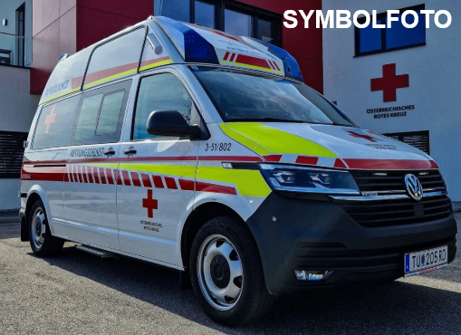 Ein weißer und roter Krankenwagen mit roten Kreuzen auf den Türen und dem Dach steht vor einem Gebäude. Er hat die Nummer 3-51/802 an der Seite. Das Kennzeichen ist TU 205 RD.