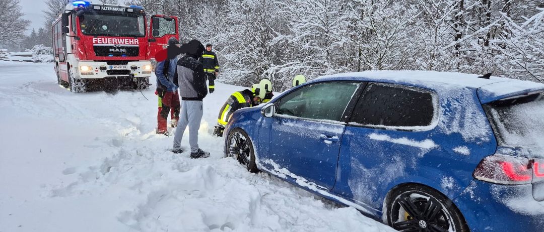 Rettungskräfte stehen um ein blaues Auto, das im Schnee stecken geblieben ist, mit einem Feuerwehrwagen im Hintergrund.