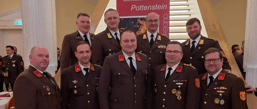 Eine Gruppe von Männern in Militäruniformen steht lächelnd auf einer Treppe, mit einem Schild hinter ihnen, das 'Pottenstein' steht. Der Hintergrund ist formell, mit einer Treppe und einer weißen Säule sichtbar.