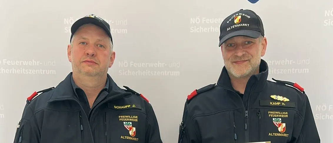 Zwei Männer in Uniform halten Auszeichnungen und stehen vor einem Hintergrund mit dem NO Feuerw Wehr Altenmarkt Logo.