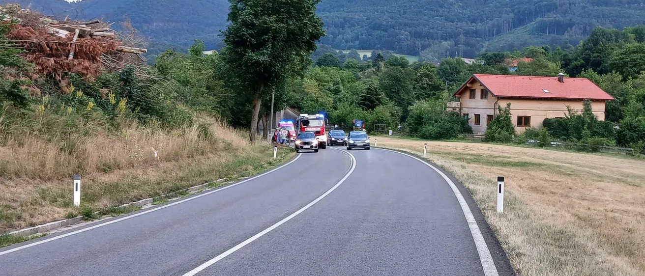 Einsatzfahrzeuge sind am Straßenrand geparkt, mit einem Haus und Bergen im Hintergrund.