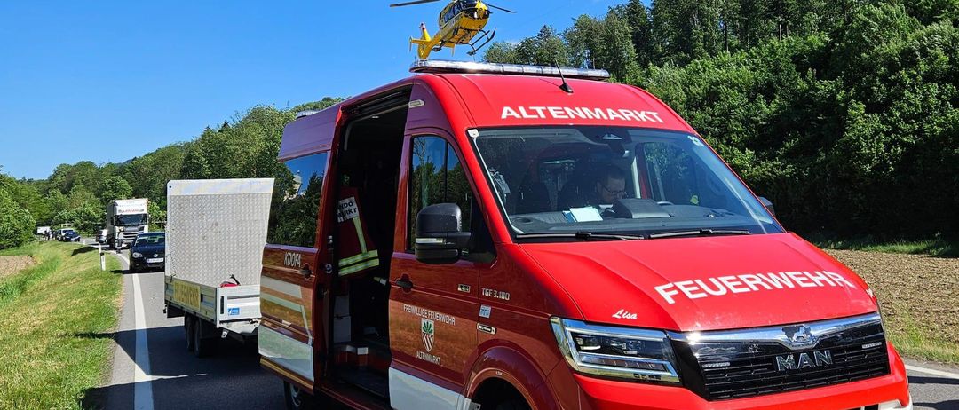 Ein roter Feuerwehrwagen mit einem Krankenwagenanhänger steht am Straßenrand, über ihm ein Hubschrauber.