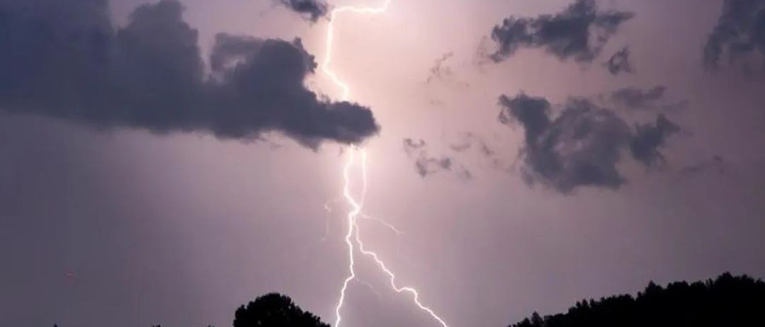 Ein Blitz schlägt mit Bäumen im Vordergrund unter dunklen, stürmischen Wolken in den Himmel ein.
