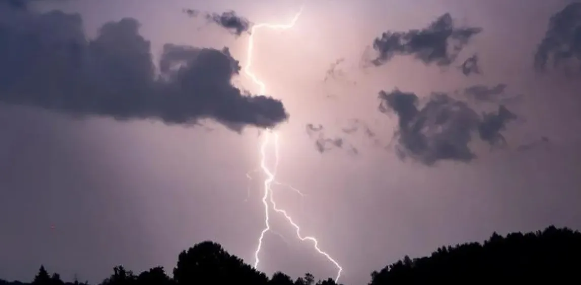 Ein Blitz schlägt mit Bäumen im Vordergrund unter dunklen, stürmischen Wolken in den Himmel ein.