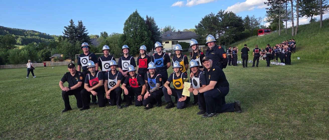Eine Gruppe von Feuerwehrleuten in Uniformen und Helmen posiert für ein Foto auf einem Feld, mit mehreren anderen im Hintergrund.
