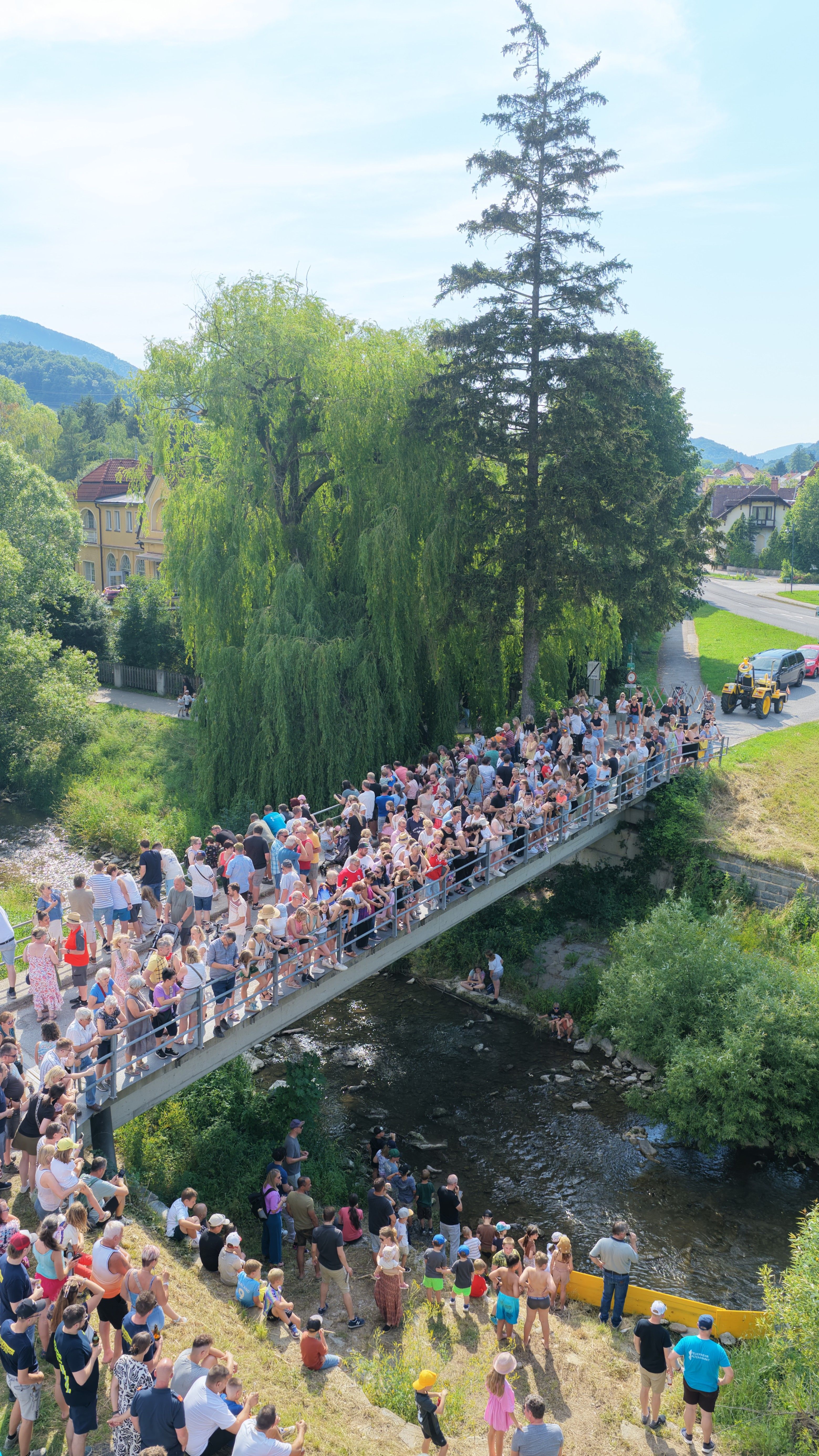 Eine große Menschenmenge versammelt sich auf einer Brücke über einem Fluss, umgeben von üppiger Vegetation. Im Hintergrund befinden sich Gebäude und Bäume, ein Fahrzeug ist auf der Straße.