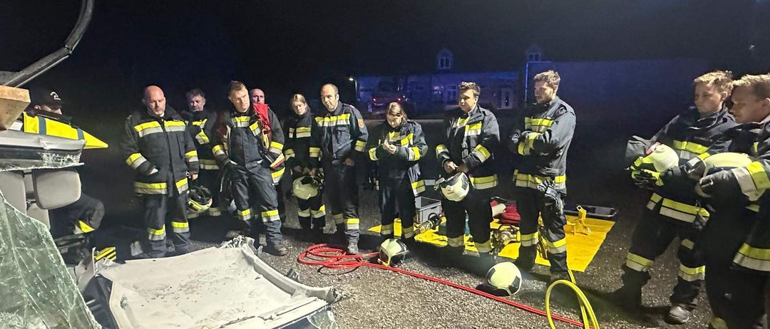 Eine Gruppe von Feuerwehrleuten steht bei Nacht um ein beschädigtes Fahrzeug herum, einige halten Helme und andere betrachten das Fahrzeug.