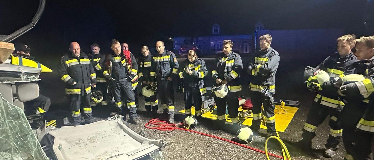 Eine Gruppe von Feuerwehrleuten steht bei Nacht um ein beschädigtes Fahrzeug herum, einige halten Helme und andere betrachten das Fahrzeug.