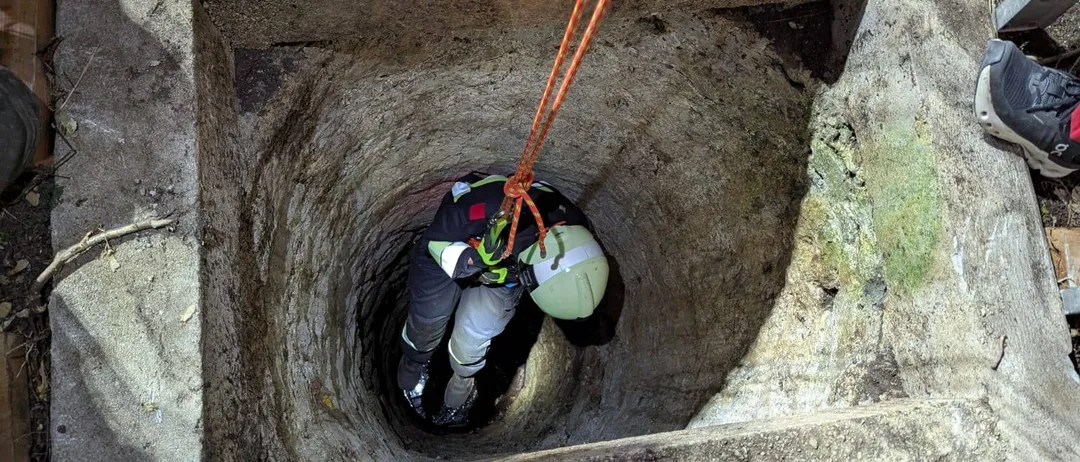 Eine Person in einer Schutzausrüstung und einem Helm steigt in einen Brunnen hinab, gesichert durch ein Seil.