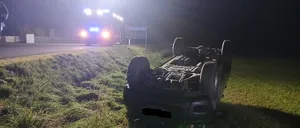 Ein Auto ist nachts auf der Straßenseite umgekippt. Die Vorderseite des Autos ist zur Straße gerichtet, die Rückseite auf dem Gras. Ein Polizeiauto ist hinter dem Auto geparkt und seine Lichter sind an.