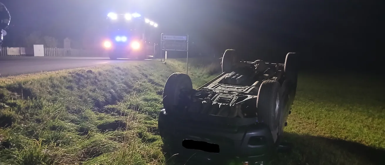 Ein Auto ist nachts auf der Straßenseite umgekippt. Die Vorderseite des Autos ist zur Straße gerichtet, die Rückseite auf dem Gras. Ein Polizeiauto ist hinter dem Auto geparkt und seine Lichter sind an.