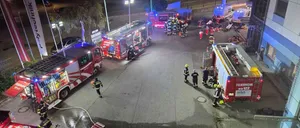 Eine Nachtaufnahme eines Feuerwehreinsatzes. Mehrere Feuerwehrfahrzeuge und Rettungswagen sind auf der Straße geparkt. Feuerwehrleute sind am Einsatzort, einige stehen herum und andere arbeiten an den Fahrzeugen.