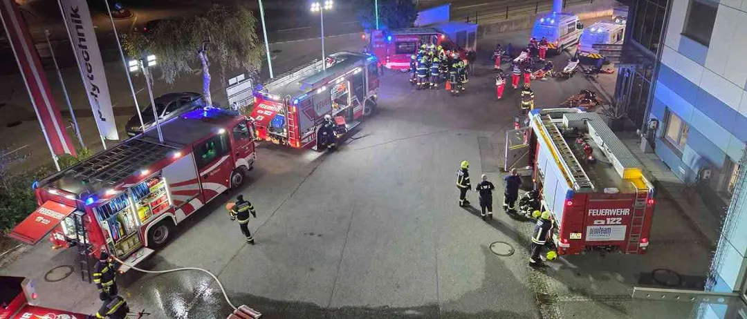 Eine Nachtaufnahme eines Feuerwehreinsatzes. Mehrere Feuerwehrfahrzeuge und Rettungswagen sind auf der Straße geparkt. Feuerwehrleute sind am Einsatzort, einige stehen herum und andere arbeiten an den Fahrzeugen.