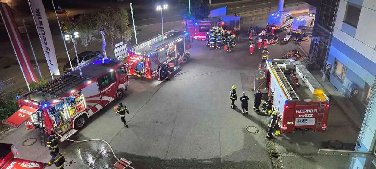 Eine Nachtaufnahme eines Feuerwehreinsatzes. Mehrere Feuerwehrfahrzeuge und Rettungswagen sind auf der Straße geparkt. Feuerwehrleute sind am Einsatzort, einige stehen herum und andere arbeiten an den Fahrzeugen.
