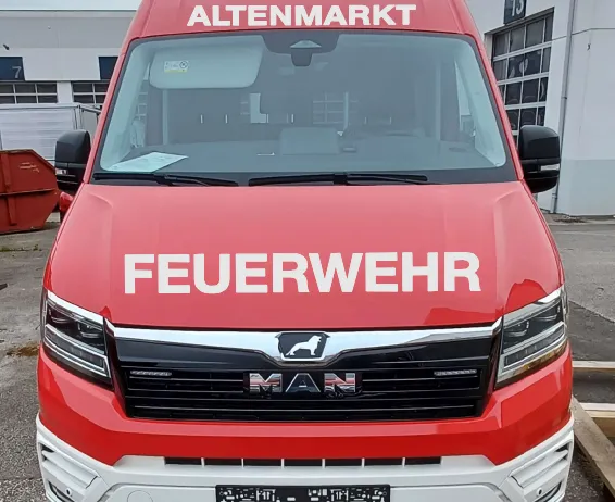 Ein roter MAN-Lkw mit den Aufschriften 'Altenmarkt' und 'Feuerwehr' steht vor einem Gebäude. Der vordere Stoßfänger hat ein weißes Design und ein schwarzes Nummernschild. Eine Nummer 13 befindet sich an der Seite des Gebäudes.