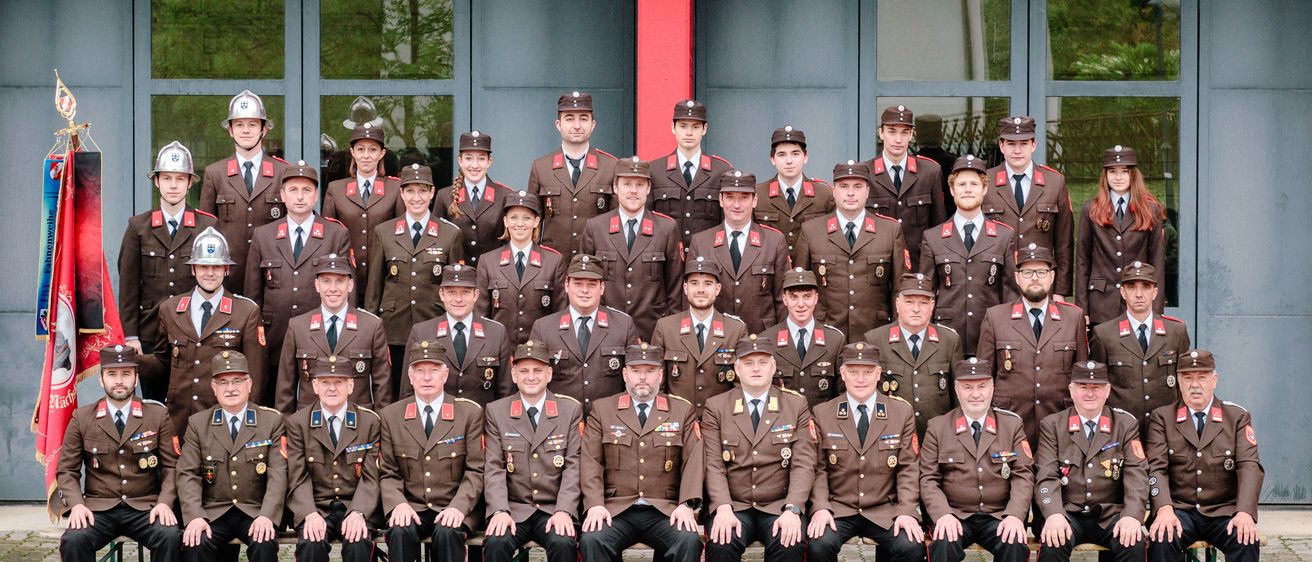 Eine Gruppe uniformierter Personen, möglicherweise Militär oder Polizei, posiert für ein Gruppenfoto. Sie tragen Hüte und Uniformen mit Abzeichen und einige haben Medaillen. Sie stehen vor einem Gebäude mit Glaswänden und einem roten Pfeiler.