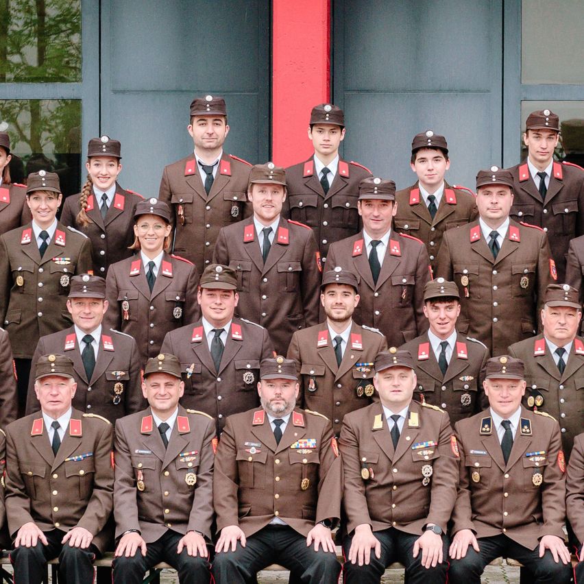 Eine Gruppe uniformierter Personen, möglicherweise Militär oder Polizei, posiert für ein Gruppenfoto. Sie tragen Hüte und Uniformen mit Abzeichen und einige haben Medaillen. Sie stehen vor einem Gebäude mit Glaswänden und einem roten Pfeiler.