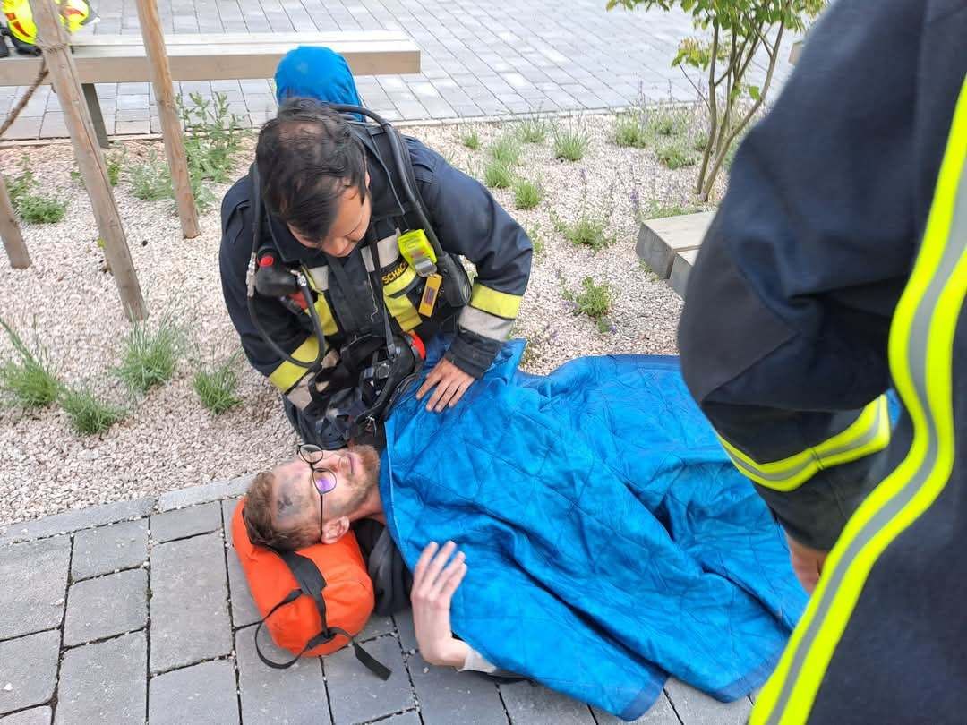 Ein Feuerwehrmann überprüft einen Mann, der auf dem Boden liegt, eine blaue Decke und eine Brille trägt, während ein anderer Feuerwehrmann in der Nähe steht und eine Bank im Hintergrund zu sehen ist.