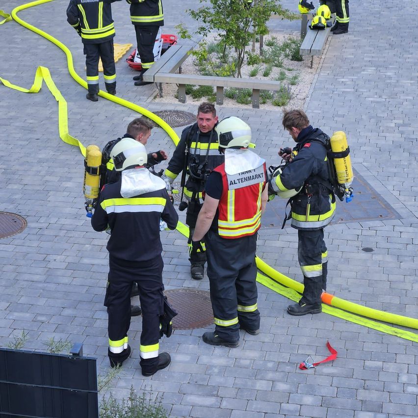 Eine Gruppe von Feuerwehrleuten in voller Ausrüstung versammelt sich auf einem gepflasterten Bereich, wahrscheinlich in Vorbereitung auf eine Brandbekämpfung. Sie sind von gelben Schläuchen umgeben.