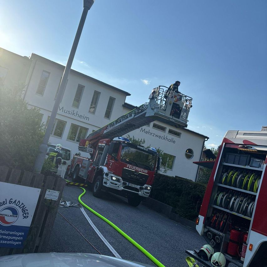 Ein Feuerwehrauto steht vor einem Gebäude mit der Aufschrift 'Musikheim'. Ein Feuerwehrmann steht auf der Leiter des Feuerwehrautos.
