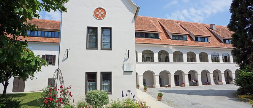 Ein weißes Gebäude mit rotem Dach, mit einem runden roten Emblem über zwei Fenstern, umgeben von einem Garten mit roten Blumen und Grün.