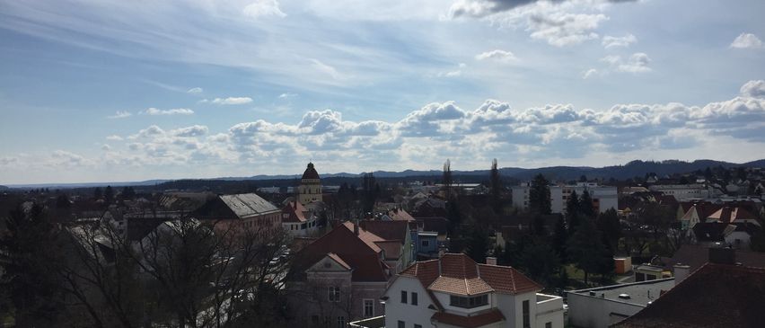 Luftaufnahme einer Stadt unter einem blauen Himmel mit verstreuten Wolken. Die Stadt hat Gebäude mit roten Dächern, von denen eines einen Turm hat. Es gibt Bäume und Autos, die vor den Gebäuden geparkt sind.