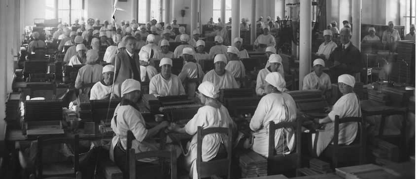 Schwarz-Weiß-Bild einer Industriefabrik mit Arbeitern in weißen Schürzen und weißen Mützen, die an Schreibtischen sitzen und an Maschinen arbeiten.