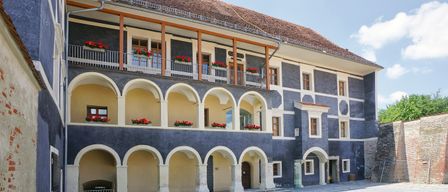 A courtyard with an archway leading to a historic building. The facade has arched windows and a balcony with red flowers.