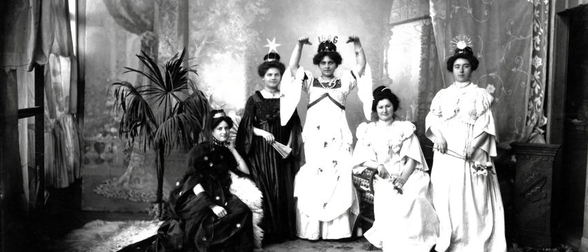 Ein Schwarz-Weiß-Foto von sechs Frauen in formeller Kleidung, zwei stehend mit erhobenen Armen und vier sitzend oder stehend um eine zentrale Figur herum, mit einem Topfpflanzen und dekorativem Hintergrund.