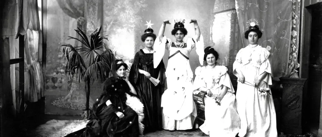 Ein Schwarz-Weiß-Foto von sechs Frauen in formeller Kleidung, zwei stehend mit erhobenen Armen und vier sitzend oder stehend um eine zentrale Figur herum, mit einem Topfpflanzen und dekorativem Hintergrund.