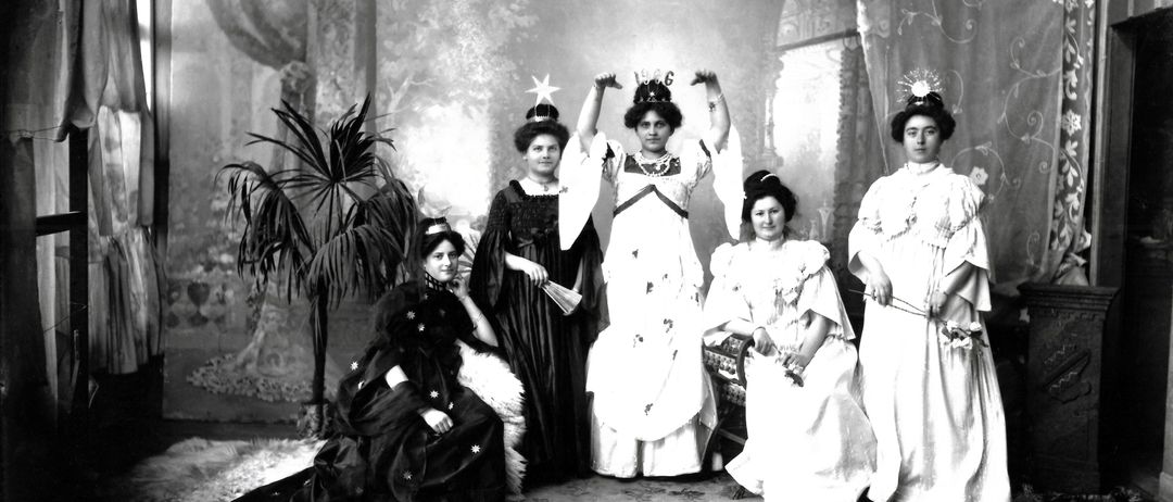 Ein Schwarz-Weiß-Foto von sechs Frauen in formeller Kleidung, zwei stehend mit erhobenen Armen und vier sitzend oder stehend um eine zentrale Figur herum, mit einem Topfpflanzen und dekorativem Hintergrund.