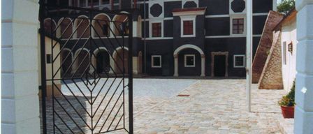 Ein schwarzes Eisentor führt in einen mit Kopfsteinen gepflasterten Innenhof. Hinter dem Tor ist ein historisches Gebäude mit Bogenfenstern und einem braunen Dach zu sehen.