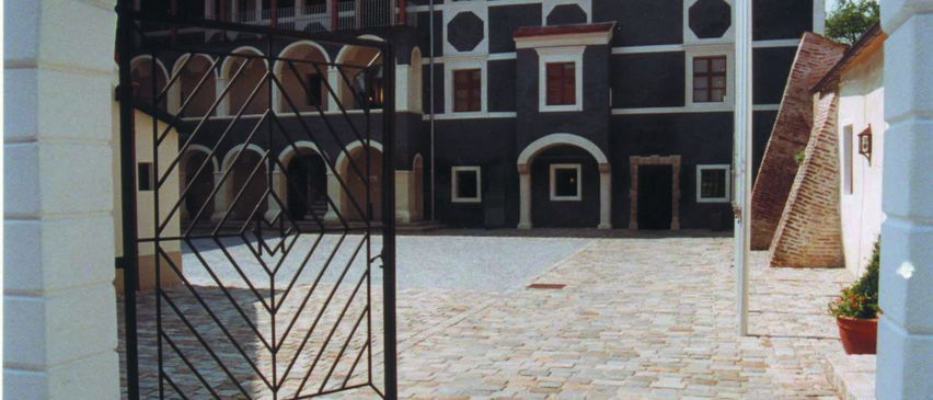 Ein schwarzes Eisentor führt in einen mit Kopfsteinen gepflasterten Innenhof. Hinter dem Tor ist ein historisches Gebäude mit Bogenfenstern und einem braunen Dach zu sehen.