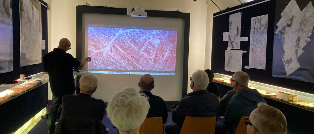 Eine Gruppe von Menschen ist drinnen versammelt und schaut sich eine Präsentation auf einer Projektionsleinwand an. Ein Mann steht davor und zeigt auf den Bildschirm. Das Publikum sitzt auf Stühlen um den Raum herum.