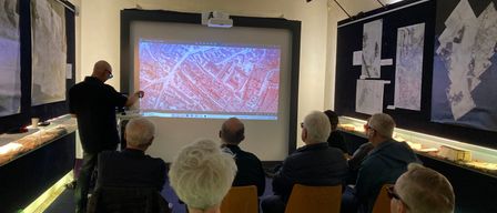 Eine Gruppe von Menschen ist drinnen versammelt und schaut sich eine Präsentation auf einer Projektionsleinwand an. Ein Mann steht davor und zeigt auf den Bildschirm. Das Publikum sitzt auf Stühlen um den Raum herum.